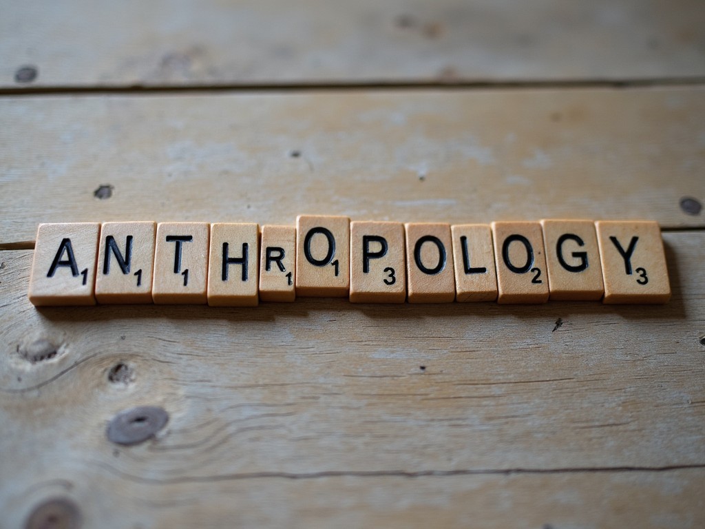 The image shows the word 'ANTHROPOLOGY' formed by wooden Scrabble tiles placed on a rustic wooden surface. The letters are clearly visible, with a mix of light and earthy tones creating a warm atmosphere. The arrangement of the tiles suggests a playful connection to education and learning. Soft natural light enhances the texture of the tiles and the wooden background. This setup invites viewers to think about the subject matter of anthropology and its importance in understanding human behavior and cultures.