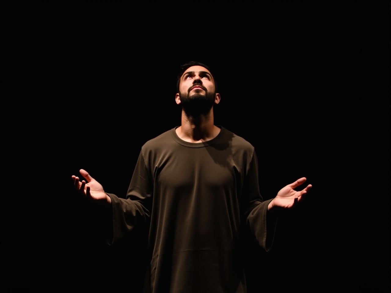 The image depicts a man in a contemplative pose, looking upward with both hands raised and open. He appears to be in a moment of prayer or deep reflection. The lighting is dramatic, creating a contrast between the dark background and the soft illumination on his face. His features are expressive, conveying a sense of hope or longing. He is dressed in a simple, flowing garment, which adds to the serene atmosphere of the scene. The overall mood is spiritual and introspective, inviting the viewer to ponder the man's thoughts.