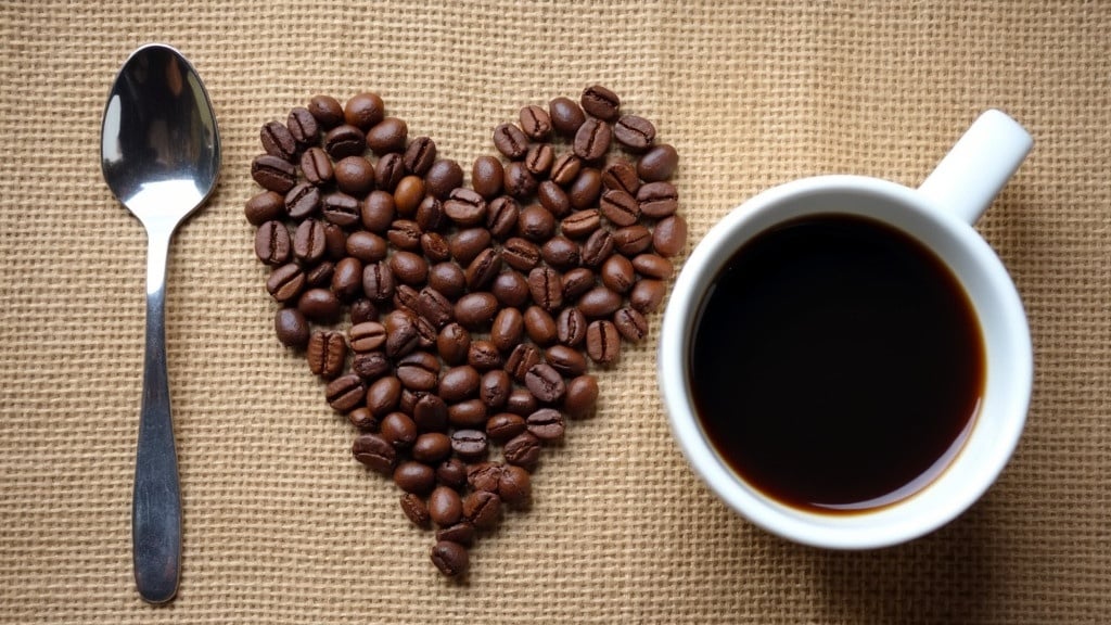 A spoon is placed on the left. Coffee beans are arranged in a heart shape at the center. A cup of black coffee is positioned on the right. The background is burlap fabric. The image conveys a love for coffee with a cozy rustic feeling. Everyday objects suggest affection for coffee. The composition is visually appealing.