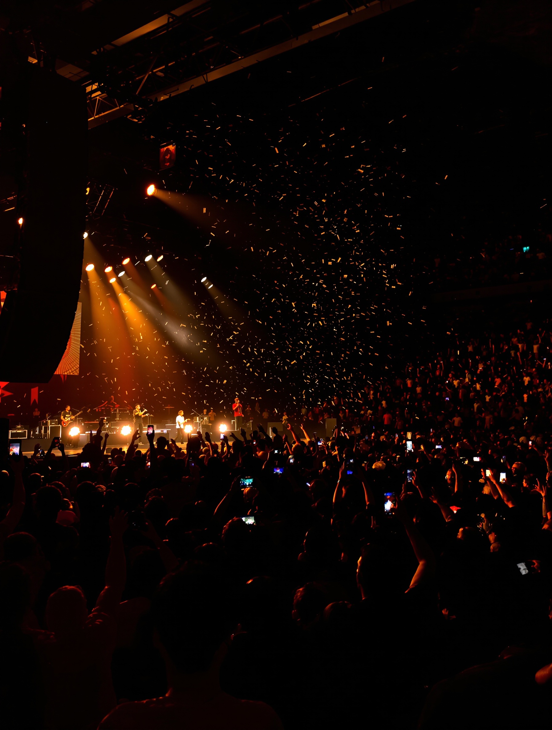 Capturing an electrifying concert atmosphere. The venue is packed with fans. Bright stage lights illuminate the performers. Energetic audience raises hands and holds phones. Confetti drifting through the air. Music resonates with loud cheer and singing.