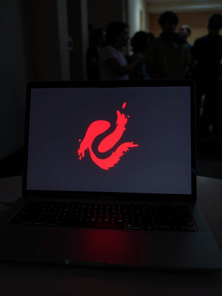 Laptop screen displays a red abstract logo. The scene is set in a dimly lit room with people visible in the background. The logo stands out against a black background.