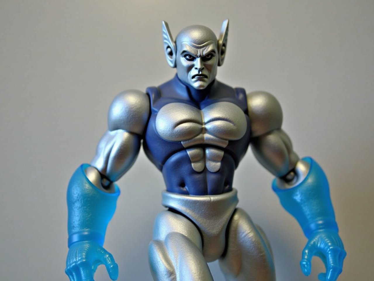 The image features a unique action figure with a sleek, metallic appearance. It has a silver and blue color scheme, with a shiny surface that catches the light. The figure has a somewhat humanoid shape, featuring exaggerated muscles and a stylized design. Its arms are equipped with translucent blue gloves that appear to glow. The character has a pointed head with distinctive features, including a subtle frown that adds to its expression. The overall design evokes a sense of futuristic or sci-fi themes, making it an eye-catching collectible.