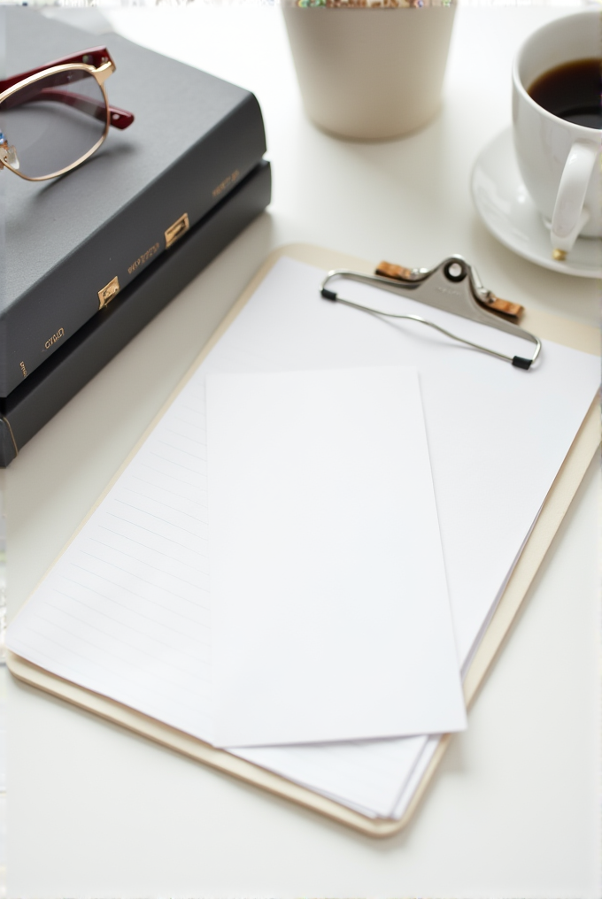 A neatly arranged desk with glasses, two stacked books, a clipboard with paper, and a cup of coffee.