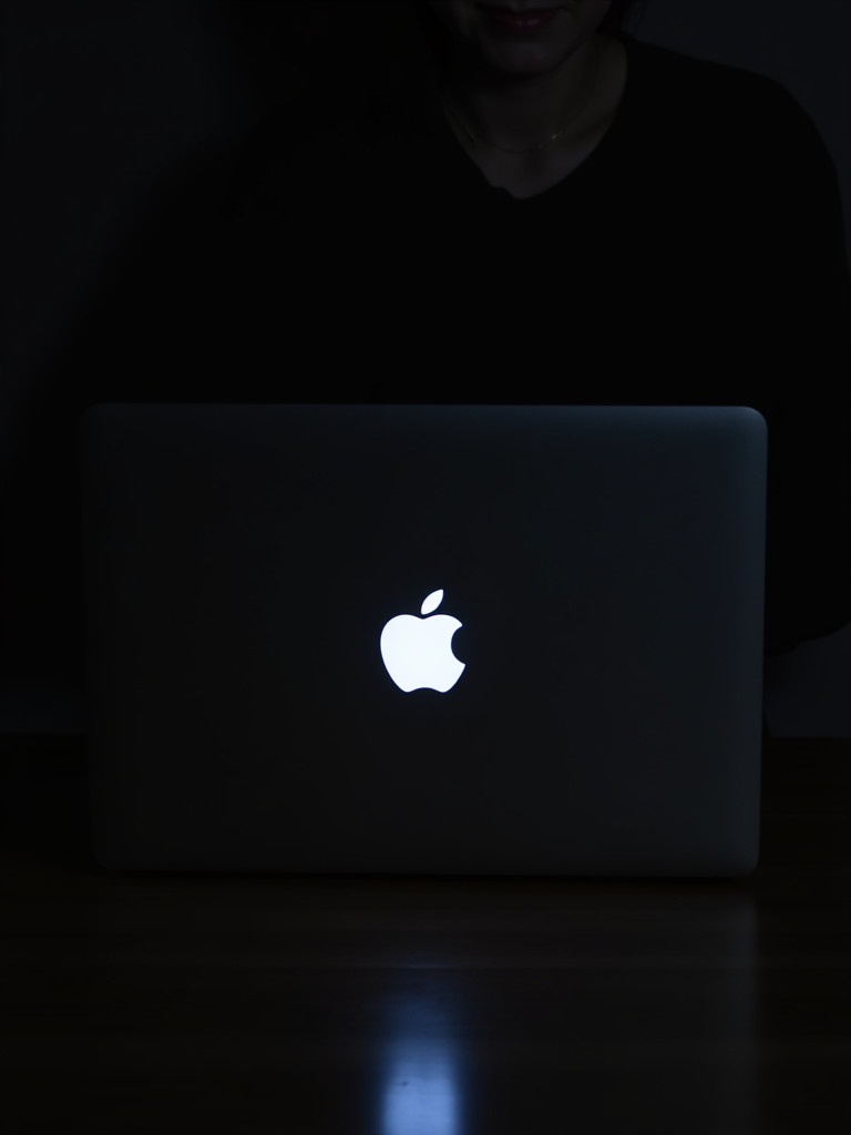 A person uses a laptop in a dark room. The Apple logo glows brightly against the darkness. The setting is intimate and focused on the screen's light.