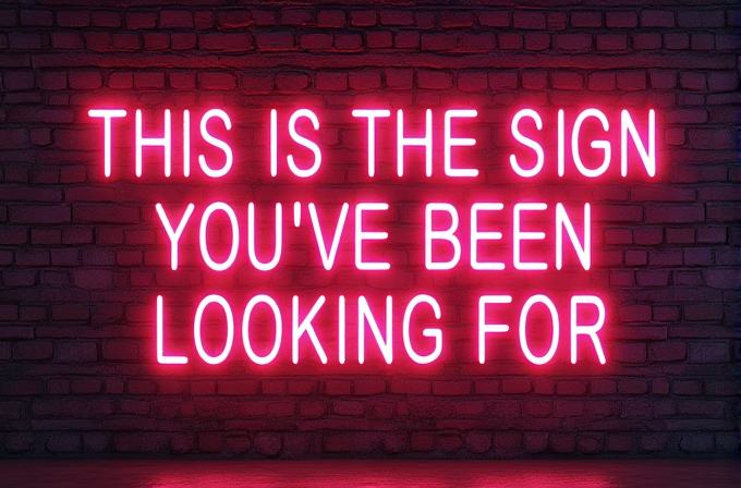 A glowing pink neon sign reads 'THIS IS THE SIGN YOU'VE BEEN LOOKING FOR' against a brick wall.