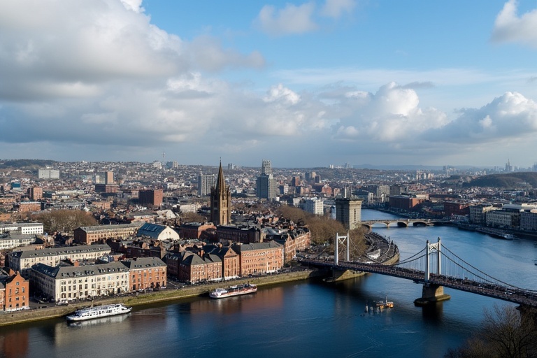 Aerial view of Dublin cityscape with bridges and River Liffey in the foreground. City skyline features historic buildings and modern structures. Clouds scatter across the blue sky.