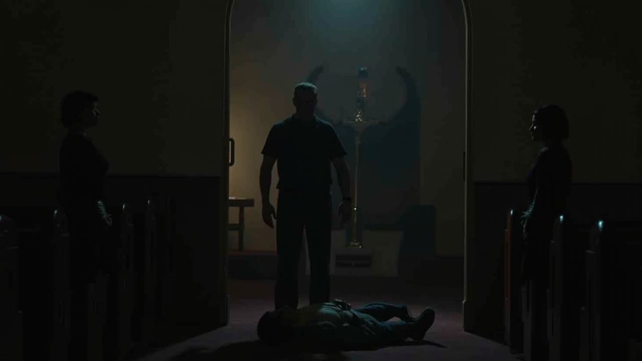 Inside a dimly lit church, a tense and dramatic scene unfolds. The husband stands over the body of a pastor, a gun still gripped in his hand. His wife remains frozen in shock, unable to comprehend the horror before her. Flickering candlelight casts long shadows, accentuating the sense of danger surrounding them. The atmosphere is thick with tragedy and suspense, making the viewer feel the weight of the moment. The church serves as a stark backdrop for this violent act, heightening the emotional stakes of the scene.