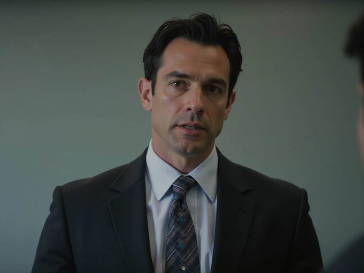 A well-dressed man in a suit and tie stands against a blurred background, suggesting an office environment. His expression is neutral, with a slight seriousness, as if he is focused or contemplating something important. The man has dark hair, neatly styled, and his attire is formal, with a dark suit jacket, light dress shirt, and patterned tie. The background is soft, with muted colors, keeping the focus on the individual. The lighting is soft and even, emphasizing his features and professional appearance.