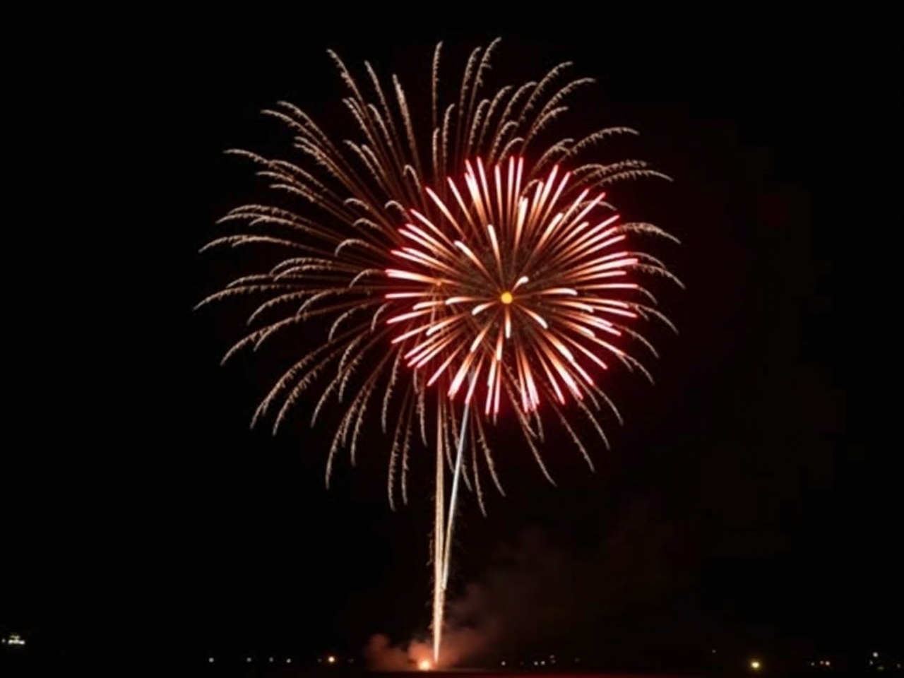 This image captures a stunning display of fireworks in the night sky, featuring a vibrant explosion against a deep black background. The colors predominantly include red, brown, and white, creating a dramatic effect. The fireworks appear to burst with energy, showcasing an ornamental halo around them. This photo, while grainy and slightly blurred due to jpeg artifacting, still evokes the excitement of festive occasions. Ideal for blogs or social media posts, it celebrates the joy of fireworks during events like the 4th of July.