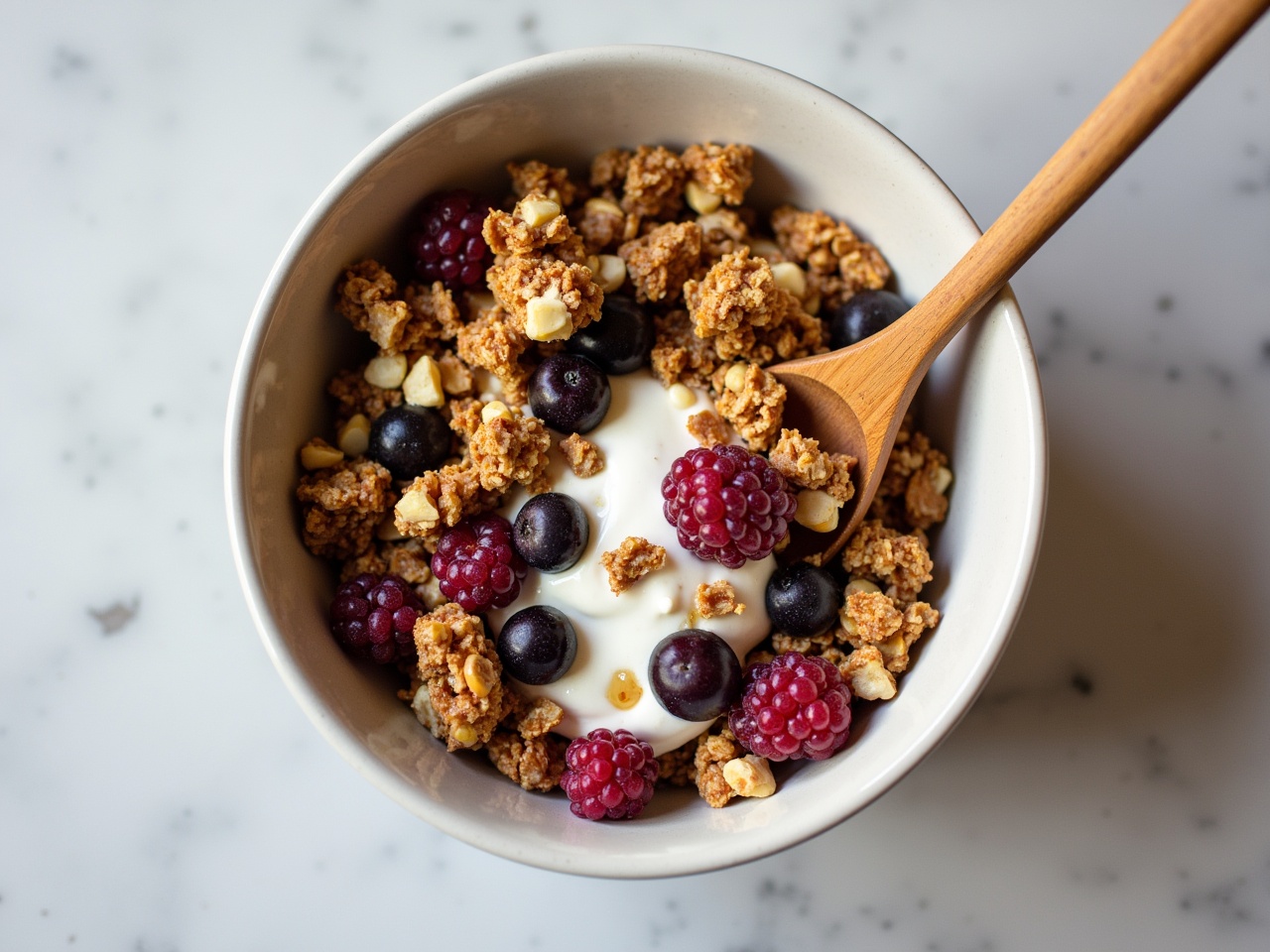 This image features a large bowl filled with delicious granola topped with creamy yogurt. Juicy berries, including blueberries and raspberries, are scattered on top. Crunchy hazelnuts add texture to the dish. A wooden spoon rests inside the bowl, ready to enjoy the meal. The background is a smooth, light-colored surface, enhancing the focus on the granola and yogurt. This visually appealing dish looks perfect for a healthy breakfast or snack.