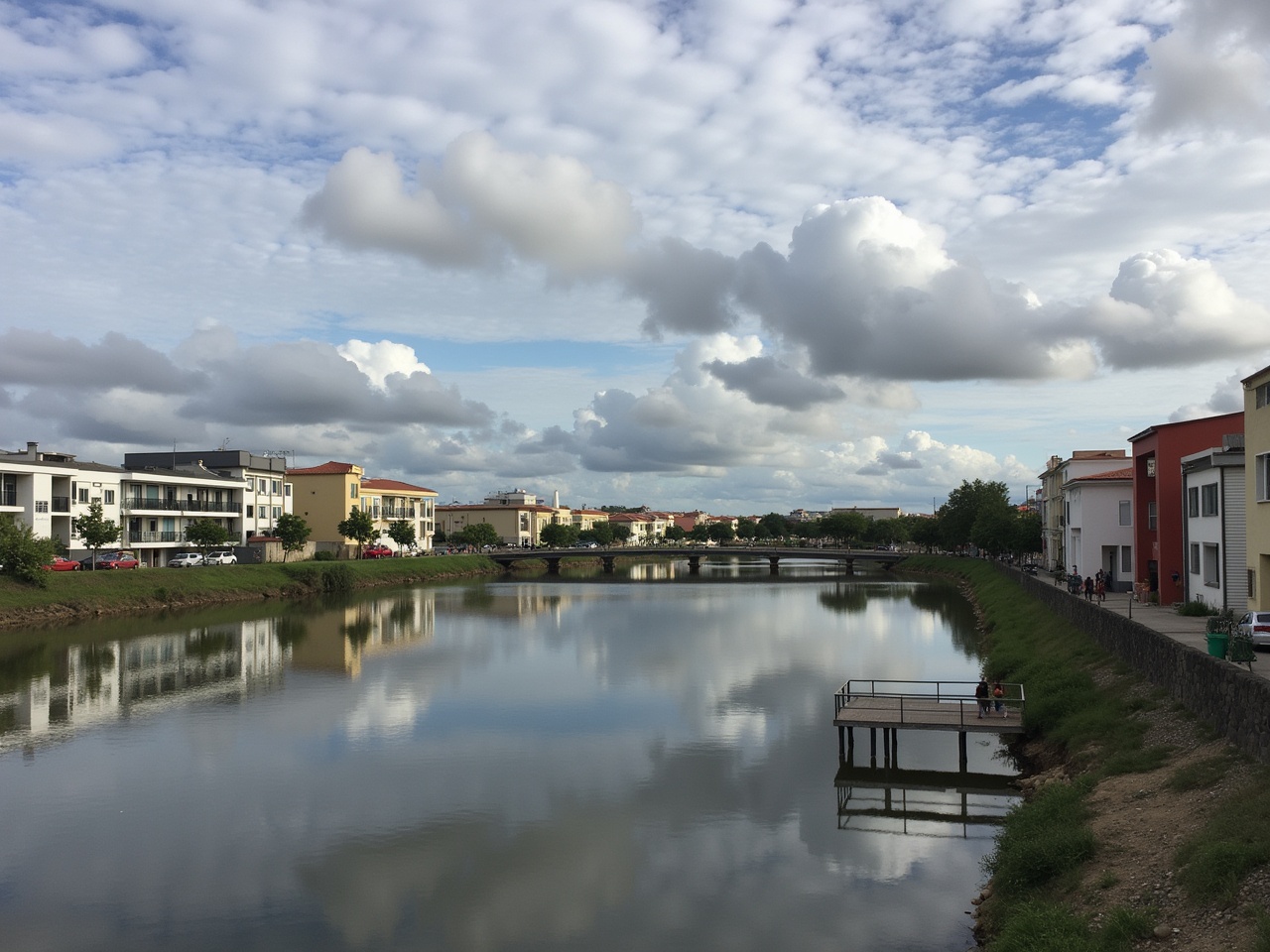 This image presents a serene urban landscape featuring a river reflecting the sky. The banks of the river are lined with modern buildings, showcasing a blend of architecture. Scattered clouds float above, casting a soft light on the water. The calm surface of the river mirrors the clouds and the structures, creating a picturesque scene. It's an ideal setting for relaxation or a leisurely stroll along the water.