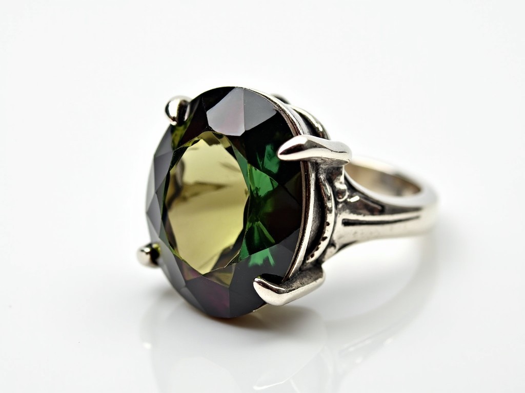 A silver ring with a large, faceted gemstone is displayed on a white background. The gemstone has a stunning array of colors, including greens, browns, and purples. The ring features a unique claw-like setting that securely holds the gemstone in place. The shiny, reflective surface of the silver complements the vibrant colors of the stone beautifully. Overall, the piece is striking and elegant, perfect for any jewelry collection.