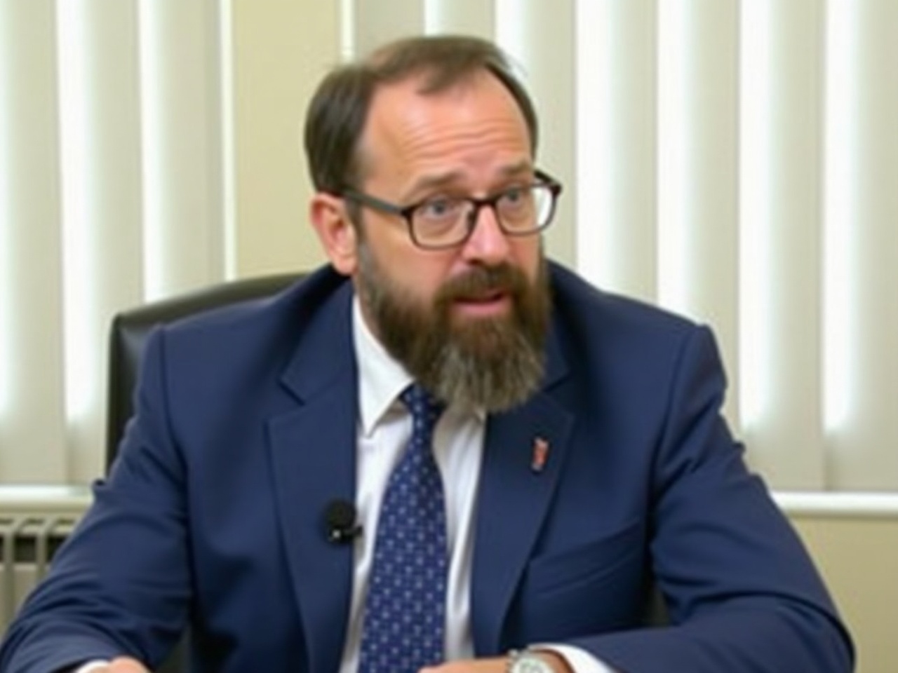 The image shows a man seated at a table. He has a well-groomed beard and a prominent mustache. The man is wearing a blue suit with a white shirt and a dotted tie. He appears to be speaking into a microphone. The background is bright and features vertical blinds, suggesting a formal setting. His glasses add to his scholarly appearance.