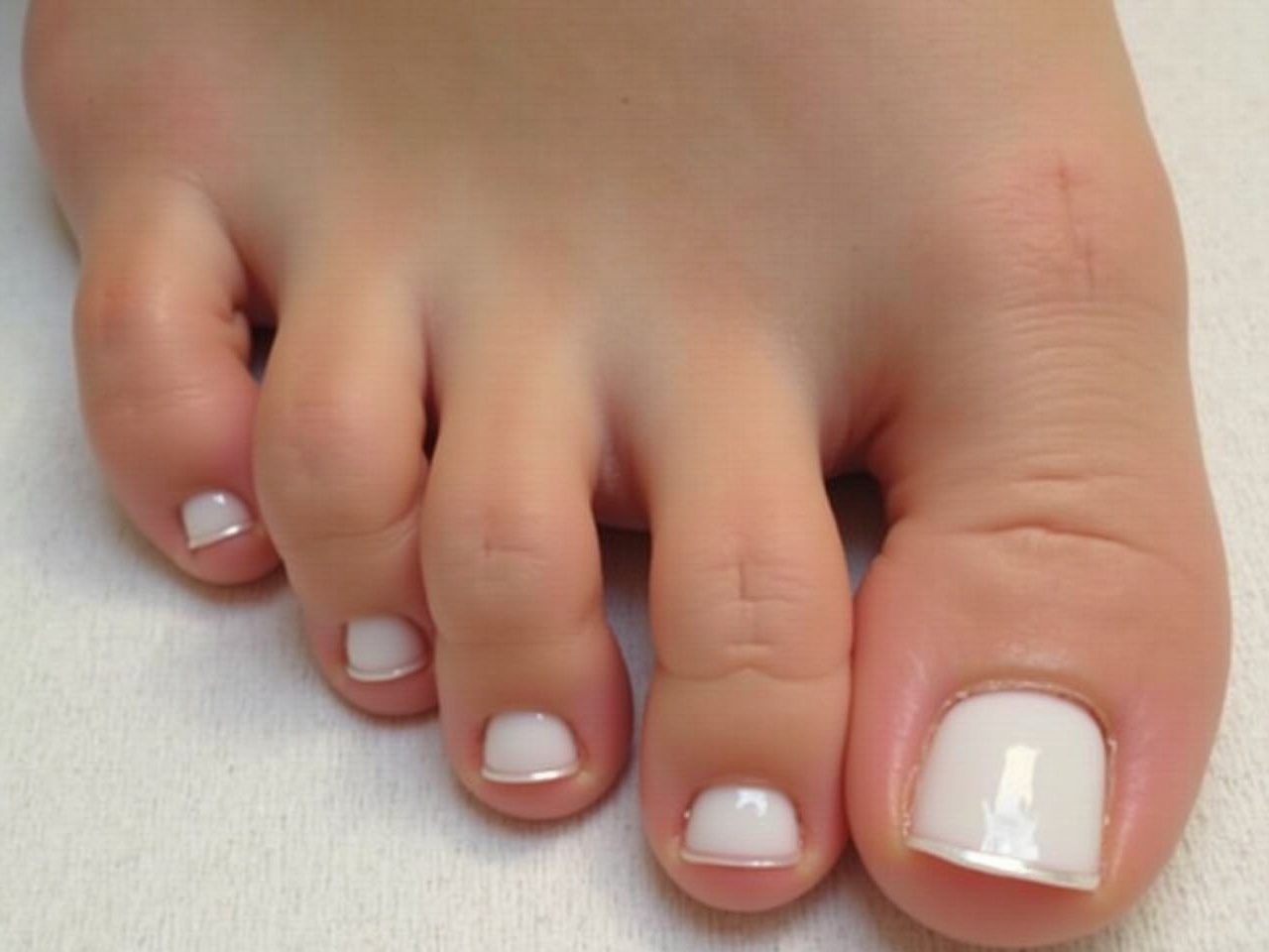 This image features a young girl's foot showcasing beautifully manicured nails. The toes are painted with a pristine white nail polish, highlighting a fresh and clean aesthetic. The skin of the foot appears smooth and well cared for, contributing to an overall impression of beauty and elegance. This close-up perspective allows for detailed observation of the neatly shaped toes and the polished nails. The lighting is soft and natural, enhancing the colors and textures visible in the image. The composition captures a tranquil and appealing view of foot grooming.