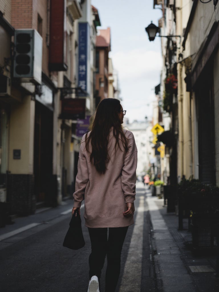 A tall woman is walking down a narrow street. The street has buildings on both sides. The woman is wearing a long pink sweatshirt. She holds a bag and walks away from the camera. It is daytime with clouds in the sky.