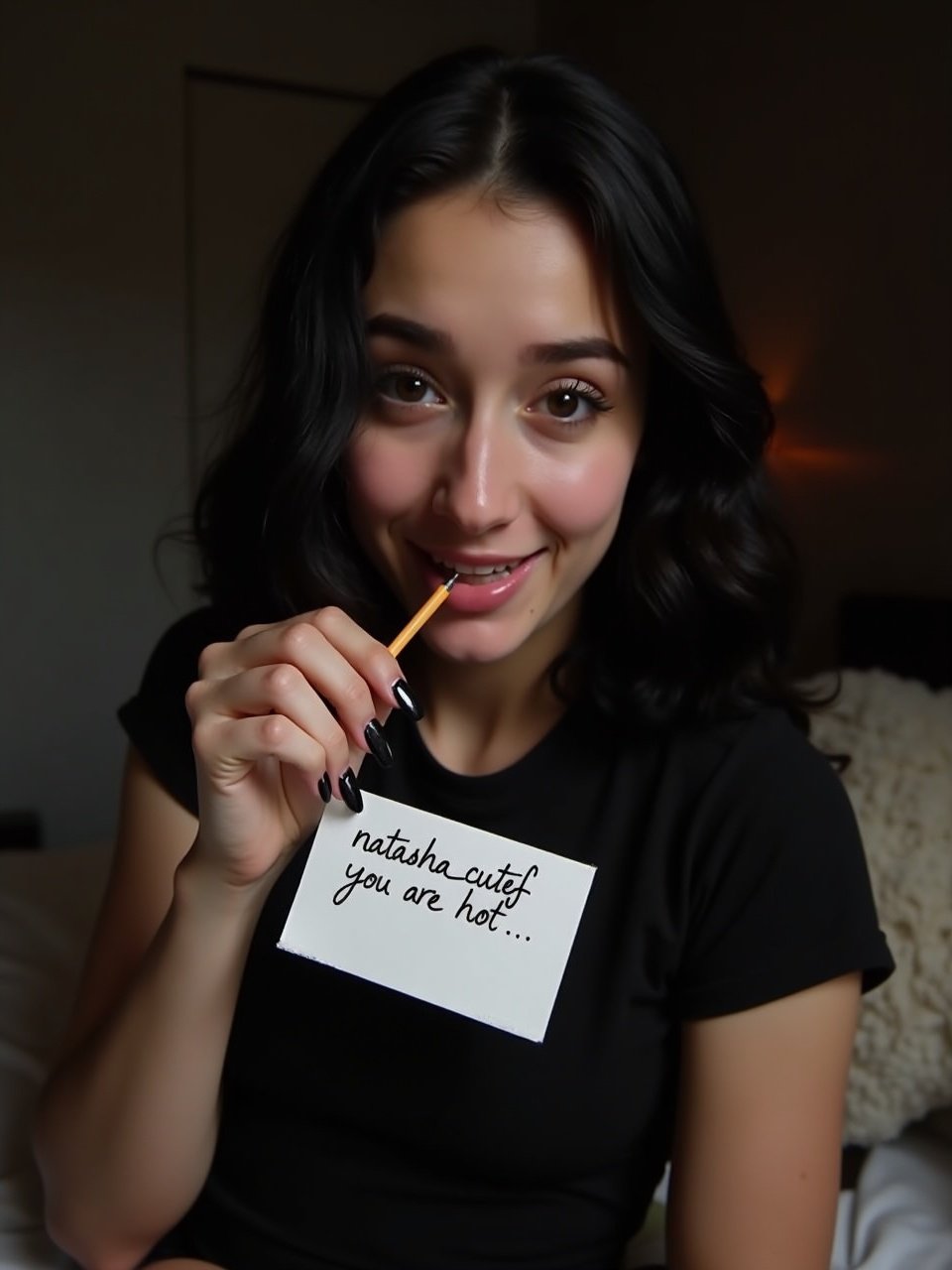 Young woman with black hair is sitting in a dark bedroom. She wears a black t-shirt and holds a piece of paper at her chest. The paper has 'natasha_cutef you are hot...' handwritten in pencil. Her nails are polished black and slightly long. She is smiling playfully and biting her lips. Her eyes show a sense of naughtiness. The lighting creates a warm atmosphere. The bedroom feels dark and inviting. This image represents a mix of intimacy and sexiness.