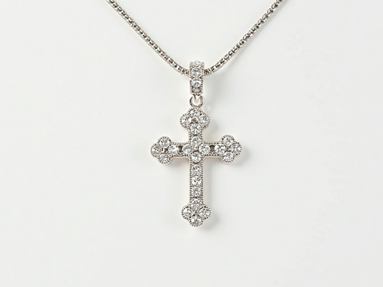 This image features a sparkling cross pendant, beautifully designed and set with numerous shiny stones. The cross is silver in color and has a polished appearance, reflecting light in a captivating way. It is suspended from a delicate chain made of similar silver material, enhancing its elegance. The overall look is one of sophistication and grace, making it suitable for both everyday wear and special occasions. The background is simple, keeping the focus on the pendant itself.