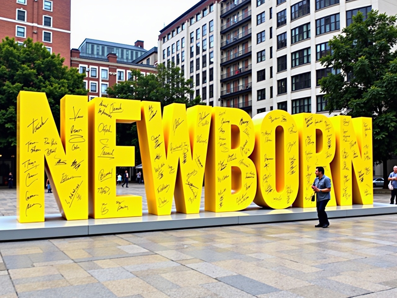 The image features a large, bold sculpture that spells out the word "NEWBORN" in bright yellow letters. The letters are positioned prominently in an outdoor area, likely a public square or plaza. Surrounding the sculpture, there are several buildings, indicating an urban setting. The letters appear to have various signatures and messages written on them, suggesting they hold cultural or commemorative significance. The scene is well-lit, indicating that the photo was taken during the day. The artwork represents a moment of celebration or remembrance, capturing the attention of passersby.