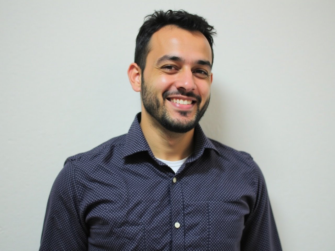 This photo features a young man dressed in a button-up shirt with a subtle pattern. He has a neat beard and is smiling broadly, giving off a friendly and approachable vibe. The background is plain, which helps focus attention on the subject. His hair is styled casually, and he is looking directly at the camera. This image captures a positive and professional appearance suitable for various applications.