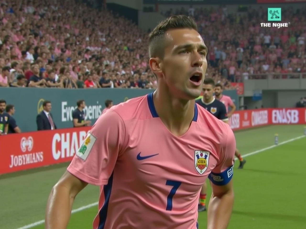 This image features a male soccer player wearing a pink jersey with the number 7. He appears to be actively engaged in the game, showing emotion as he plays. The background is filled with a cheering crowd, indicating a lively soccer match. The player is the focal point, emphasized by his close positioning in the frame. The atmosphere is electric, typical of a major sporting event. This moment captures the essence of competitive spirit in soccer.