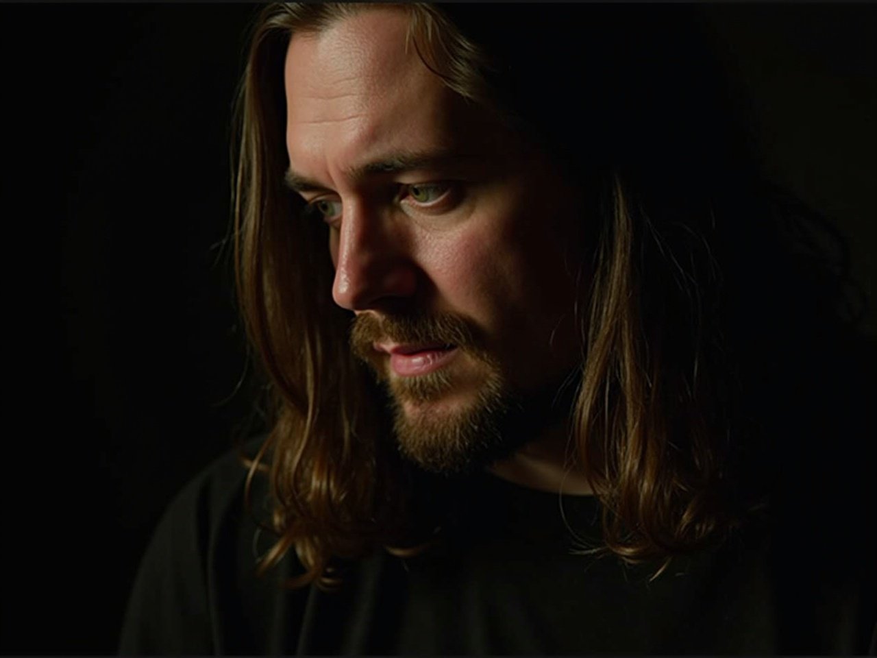 The image features a close-up portrait of a man with long hair. His expression is serious and contemplative, as he looks slightly downward. His face is adorned with facial hair, giving him a rugged appearance. The lighting highlights the texture of his skin and the strands of his hair. The background is dark, emphasizing the subject's features. The overall mood is reflective and intense.