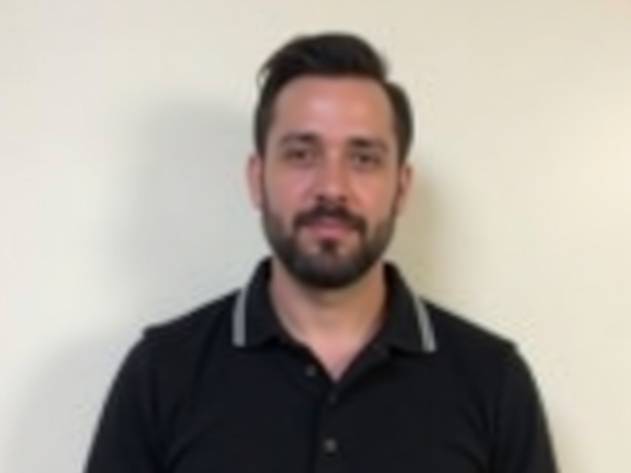 The image shows a man with dark hair and a beard, wearing a black polo shirt with a light stripe around the collar. He is standing in front of a plain white wall, and he is looking directly at the camera with a neutral expression on his face. The lighting appears to be soft, highlighting the features of his face effectively. His hair is styled back, and he has a relatively even complexion. There are no visible accessories or objects in the image, keeping the focus on the individual.