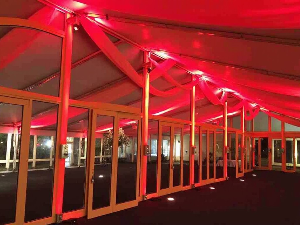 The image shows a spacious event tent with a bold red lighting scheme. The ceiling and walls are adorned with soft red fabric, creating a warm and inviting atmosphere. The floor is dark, emphasizing the illuminated features. Large glass doors provide access to additional spaces or outdoor views. This venue is ideal for various events, including weddings and corporate parties.