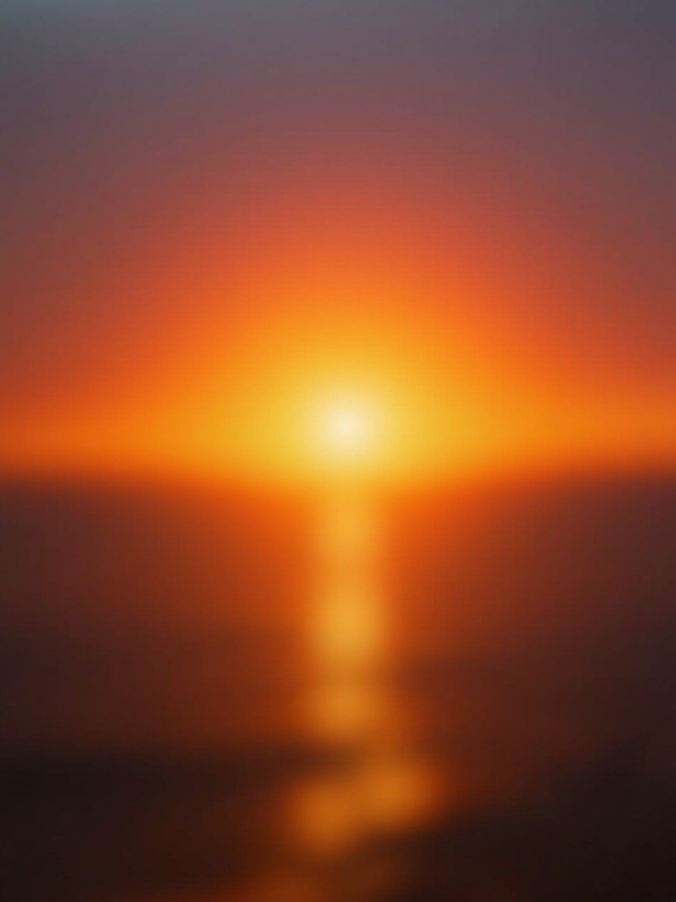 A warm and blurred image of a setting sun over a calm ocean, casting a vivid reflection on the water. The colors transition from deep oranges and reds at the center, fading into purples and blues at the edges, creating a soothing, impressionistic view of a sunset.