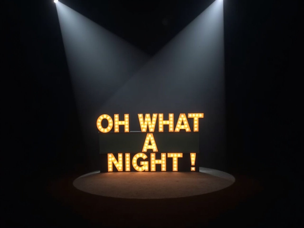 A spotlight casts dramatic beams across the dark background, illuminating a sign reading 'OH WHAT A NIGHT!' in yellow marquee bulb lights. The atmosphere is filled with excitement and anticipation. The sign stands as the focal point in the scene, drawing attention with its bright lights. This setup suggests a lively event or performance is about to take place. A sense of grandeur is conveyed by the spotlight and arrangement.