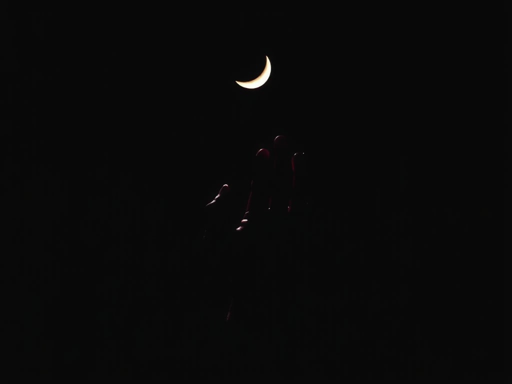 A silhouetted hand reaching towards a crescent moon in a dark sky.