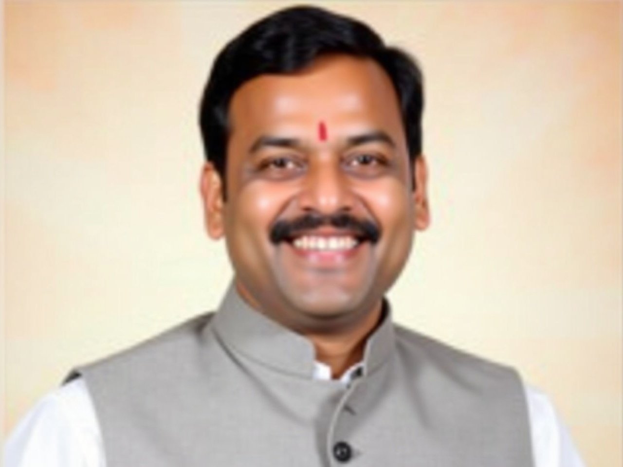 The image shows a man with a friendly smile, wearing a traditional Indian outfit. He has a light gray vest over a white shirt. There is a small red mark on his forehead, which is common in some Indian cultures. The background appears to be a soft gradient of colors, contributing to a formal yet warm atmosphere. The man appears to be in a posed setting, likely for a professional or ceremonial purpose.